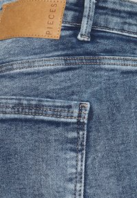 Blå denimjeans med en läderetikett vid midjan. Tyget har en blekt textur med synliga sömmar och en bakficka.