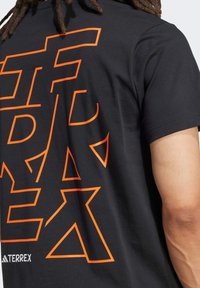 Camiseta de algodón negra con gran texto geométrico naranja en la espalda. Incluye un detalle de logo blanco cerca del dobladillo. Textura suave, corte regular.