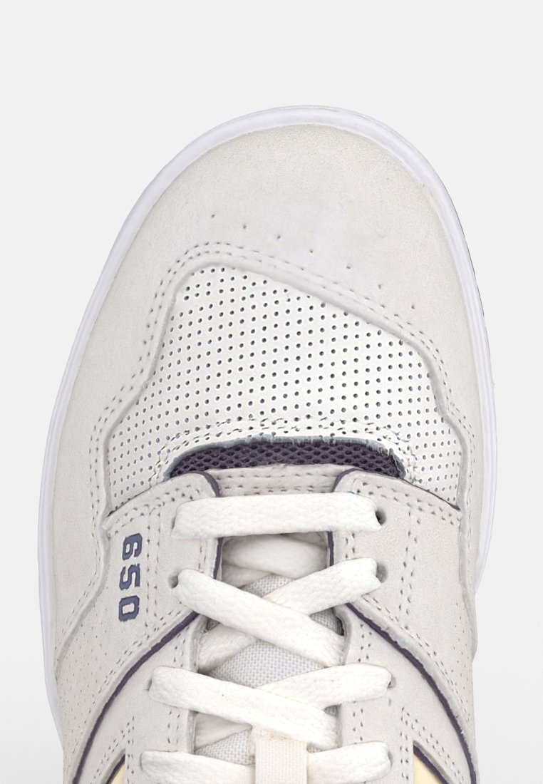 Sneaker bianco con tomaia in suede perforato, dettagli in contrasto viola, lacci piatti e suola in gomma. Il numero di stile "650" è visibile.