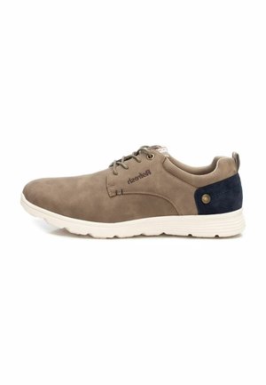 Zapatilla de ante marrón con suela de goma blanca, forma redondeada y acento azul marino en el talón. Presenta un diseño de cordones y una marca en el lateral.