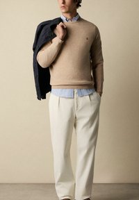 Maglione beige a maglia con scollo rotondo, indossato sopra una camicia colletto azzurra, abbinato a pantaloni a coste crema e una giacca blu navy.