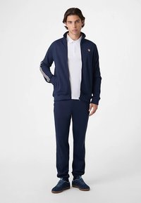 Marineblauw trainingspak met witte streepaccenten, ritsjack, lange mouwen en aangesloten broek. Combineer met blauwe textuursneakers voor een casual look.