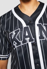 Černý baseballový dres s bílými pruhy, velkým nápisem "KAN" a designem s V výstřihem s kontrastními bílými lemy a černými knoflíky.