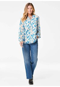 Blaue und weiße gemusterte Bluse mit langen Ärmeln und rundem Ausschnitt, kombiniert mit hochgeschlossenem Jeans und schwarzen Stiefeletten.