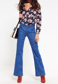 Jeans blu a campana con vita aderente, abbinati a una blusa a fiori blu navy e stivaletti rossi alla caviglia. Include una borsa bianca e nera.