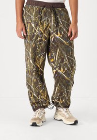 Camouflage sweatpants with a brown, yellow, and green pattern, featuring an elastic waistband and adjustable cuffs, paired with beige sneakers.