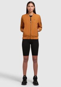 Bronzen bomberjack met ritssluiting, geribbelde manchetten en zijzakken, gecombineerd met zwarte fietsbroek en zwarte sneakers. Eenvoudig ontwerp.