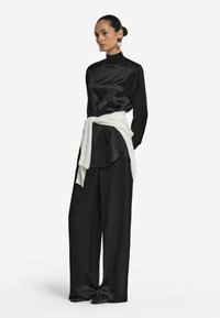 Schwarze Satinbluse mit hohem Kragen, kombiniert mit lockeren schwarzen Hosen. Weißer Pullover, der um die Taille gebunden ist, spitze schwarze Schuhe.