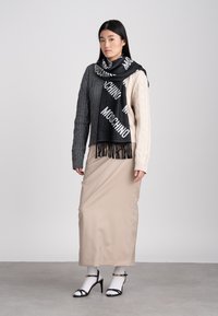 Kvinna iförd en tvåtonad kabelstickad tröja, svart Moschino-scarf, beige långkjol, vita strumpor och svarta sandaletter, stående på ett grått golv.
