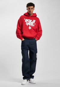 Felpa rossa con testo grafico bianco, abbinata a pantaloni cargo blu scuro con tasche laterali e scarpe da ginnastica bianche. Insieme semplice e casual.
