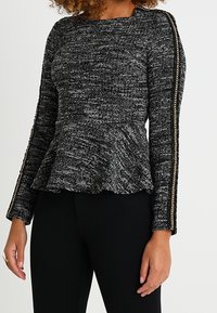 Top peplum a maniche lunghe con texture in bianco e nero, caratterizzato da uno scollo quadrato e dettagli metallici lungo i bordi delle maniche.