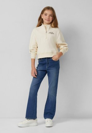 Jong meisje staat met een hand in de zak, draagt een crèmekleurige ritsvest, blauwe jeans en witte sneakers tegen een effen achtergrond.