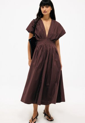 Kvinde iført en brun midi-kjole med dyb V-udskæring, korte ærmer og læg, bærende en sort flet taske og iført sorte hælede sandaler.