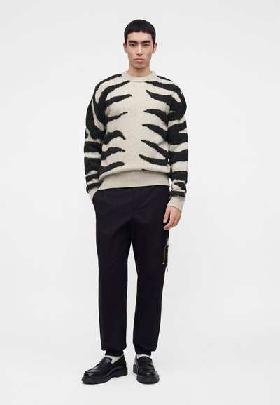 Homme debout portant un pull beige et noir à rayures tigre, pantalon noir avec une poche latérale zippée, chaussettes blanches et mocassins noirs.
