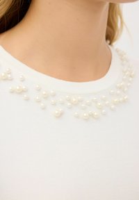 Vit stickad topp med rund halsringning prydd med kluster av små, runda, konstgjorda pärlor i varierande storlekar längs kragen.