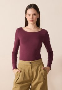 Langermet, tettsittende topp i dyp plommefarge med vid scoop-hals, kombinert med høytlivet, plisserte khaki-bukser med en knapp.