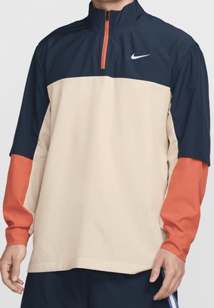 Mand iført en Nike pullover med marineblå, beige og orange farveblokke og en kort lynlås foran ved kraven.