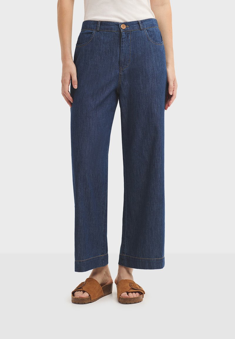 Pantalon en denim coupe large de couleur bleu foncé, avec poches avant, fermeture par bouton et lignes épurées. Associé à des sandales marron à enfiler.