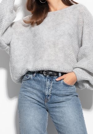 Femme portant un pull en grosse maille gris clair rentré dans un jean bleu taille haute avec une ceinture noire, main dans la poche.