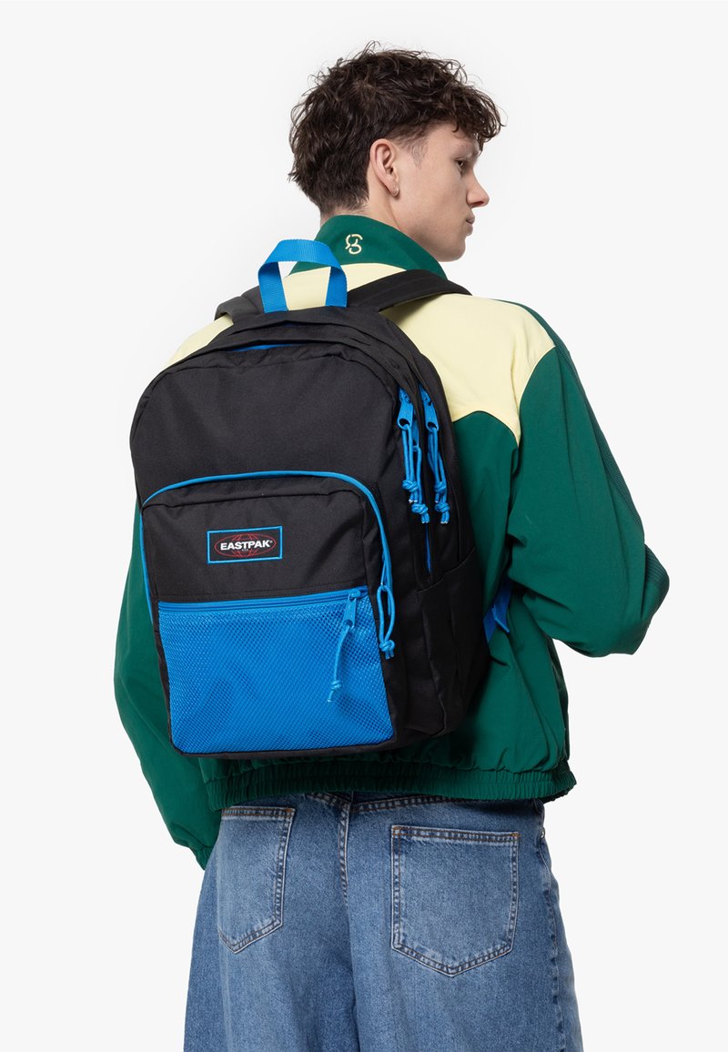 Mochila Eastpak negra con acentos azules, presente un bolsillo de malla en la parte delantera y tirantes ajustables, llevada en la espalda de una persona.