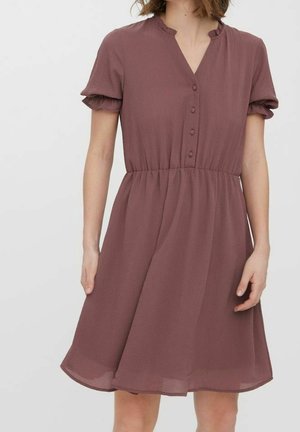 Vrouw die een knielange mauve jurk draagt met korte pofmouwen, een V-hals met knopen en een gerimpelde taille, tegen een effen lichtgrijze achtergrond.