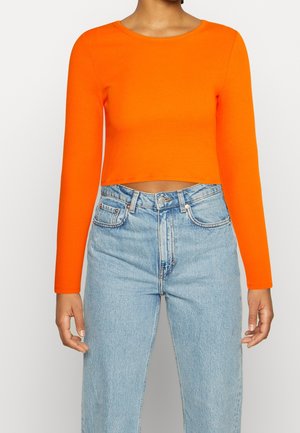 Vrouw draagt een feloranje cropped top met lange mouwen, gecombineerd met een hoogwater lichtblauwe denim jeans, tegen een effen achtergrond.