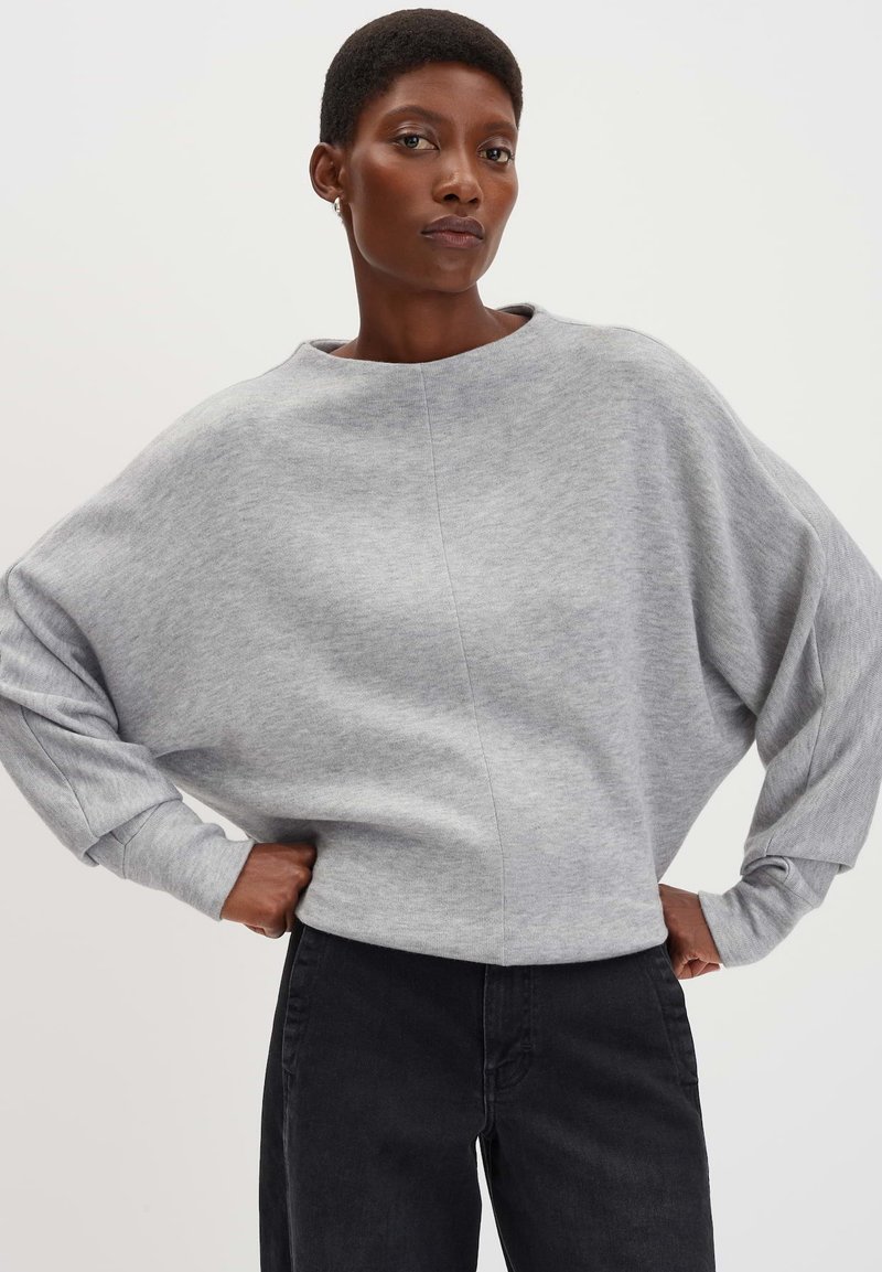 Felpa grigia con ampia scollatura a barca, vestibilità rilassata e maniche lunghe. Abbinata a jeans neri, che mostrano uno stile casual e confortevole.