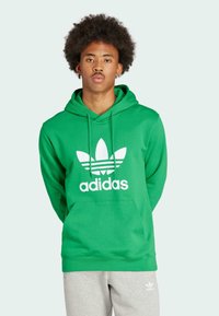 Sudadera de algodón verde con un gran logo blanco de Adidas y diseño de tres rayas, que cuenta con un bolsillo tipo canguro en la parte delantera y capucha ajustable.