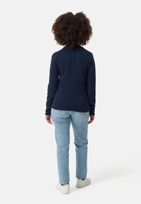 Marineblauer Strickpullover mit langen Ärmeln, strukturiertem Stoff und tailliertem Design, kombiniert mit hellblauen Jeans und weißen Turnschuhen.