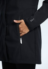 Veste noire imperméable en tissu doux, avec une poche zippée et des poignets ajustables, au texture lisse et au design élégant.