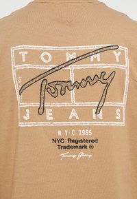 Beige särk, mille tagaküljel on valge ääristatud ruudustik, millel on suur must kiri "Tommy" ja tekst "TOMMY JEANS NYC 1985 NYC registreeritud kaubamärk".