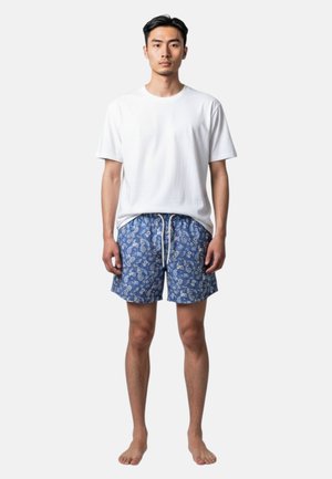 Giovane uomo in piedi a piedi nudi, indossa una t-shirt bianca semplice e pantaloncini blu con motivo floreale e coulisse bianca.