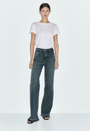 White t-shirt, loose fit, paired with dark wash wide-leg jeans. Flat sandals with a fringed hem detail, solid background.