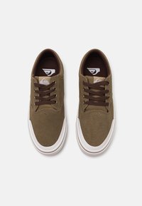 Brune sued sneakers med hvite gummisåler, med brune snørebånd og en logobakgrunn på tungen. Rundet tådesign og perforerte detaljer.