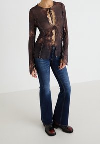 Haut marron transparent avec bords festonnés, détails en lacets et décolleté en V profond. Associé à un jean bleu évasé et des bottines noires.