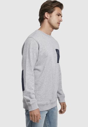 Fiatal férfi jobbra néz, világosszürke pulóvert visel, sötétkék cipzáras mellzsebbel és hozzáillő könyökfolttal, világoskék farmerrel párosítva.