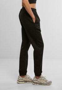 Pantalon de survêtement en coton noir avec taille élastique et poignets, doté de poches latérales. Assorti à des chaussures de sport beige et grises.