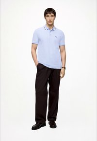 Homme portant un polo Boss bleu clair, un pantalon noir, des chaussures foncées et une montre-bracelet, debout avec une main dans la poche sur fond blanc.