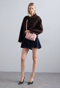 Femeie îmbrăcată cu o jachetă supradimensionată maro închis, fustă bleumarin, tocuri negre și ținând o geantă de designer roz cu curea de umăr.