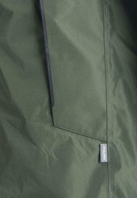 Tessuto della giacca verde scuro con una superficie liscia. Presenta una tasca laterale e una piccola etichetta con la dicitura "JACK & JONES". Design pulito e minimalista.
