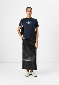 Námornícke bavlnené tričko s bielym a svetlomodrým logom Calvin Klein, skombinované s čiernou koženou maxi sukňou a krémovými slip-on topánkami.