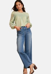 Blusa verde chiaro con dettagli in pizzo, jeans in denim blu a vita alta e sandali con cinturini marroni, dotati di tasche con cuciture decorative.