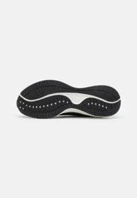 Suela de zapato deportivo negro con un patrón de banda de rodadura prominente, ranuras texturizadas y un acento de media suela blanca. Presenta bordes curvados para mayor flexibilidad.