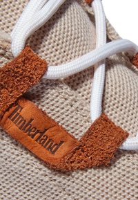 Baskets beiges en tissu texturé, lacets blancs passés à travers des accents en suède marron, avec un logo Timberland sur une étiquette en cuir.