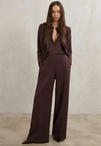 Bordó szabott öltöny széles szárú nadrággal és mély V-nyakú blúzzal. Sima anyag, minimalista dizájn, hegyes orrú magassarkú cipő.