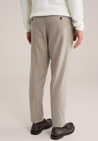 Mann in beigen, maßgeschneiderten Hosen, weißem Pullover und dunkelbraunen Lederloafers, der vor einem einfarbigen, hellen Hintergrund steht.