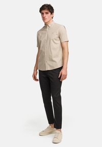Korte mouw beige shirt met een button-down kraag, gecombineerd met donker geruite broeken en beige sneakers, staand tegen een witte achtergrond.