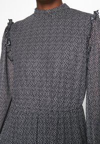 Robe noire à petits pois blancs, manches longues bouffantes en voile, col montant et détails à volants aux épaules.