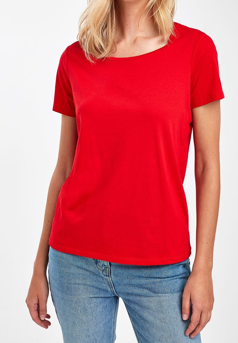Femme portant un t-shirt rouge uni à manches courtes et un jean bleu clair, debout devant un fond blanc uni.