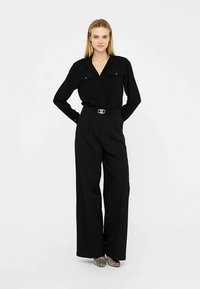 Chemise noire à manches longues avec deux poches poitrine, associée à un pantalon noir taille haute à jambes larges, équipé d'une ceinture avec boucle dorée.